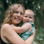A joyful mother embraces her baby outdoors in a lush green forest.